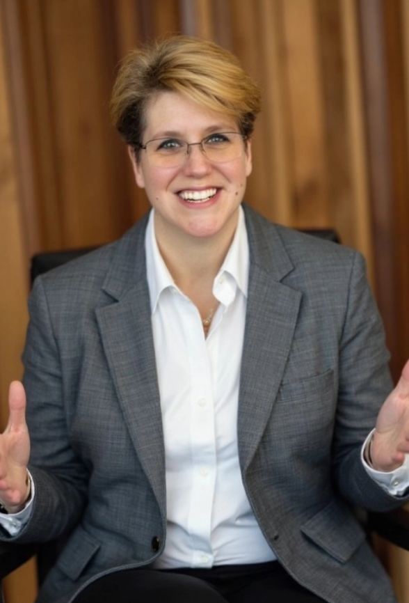 Portrait of Tara Killen smiling while seated in front of a wood-paneled background, wearing a gray blazer and white shirt